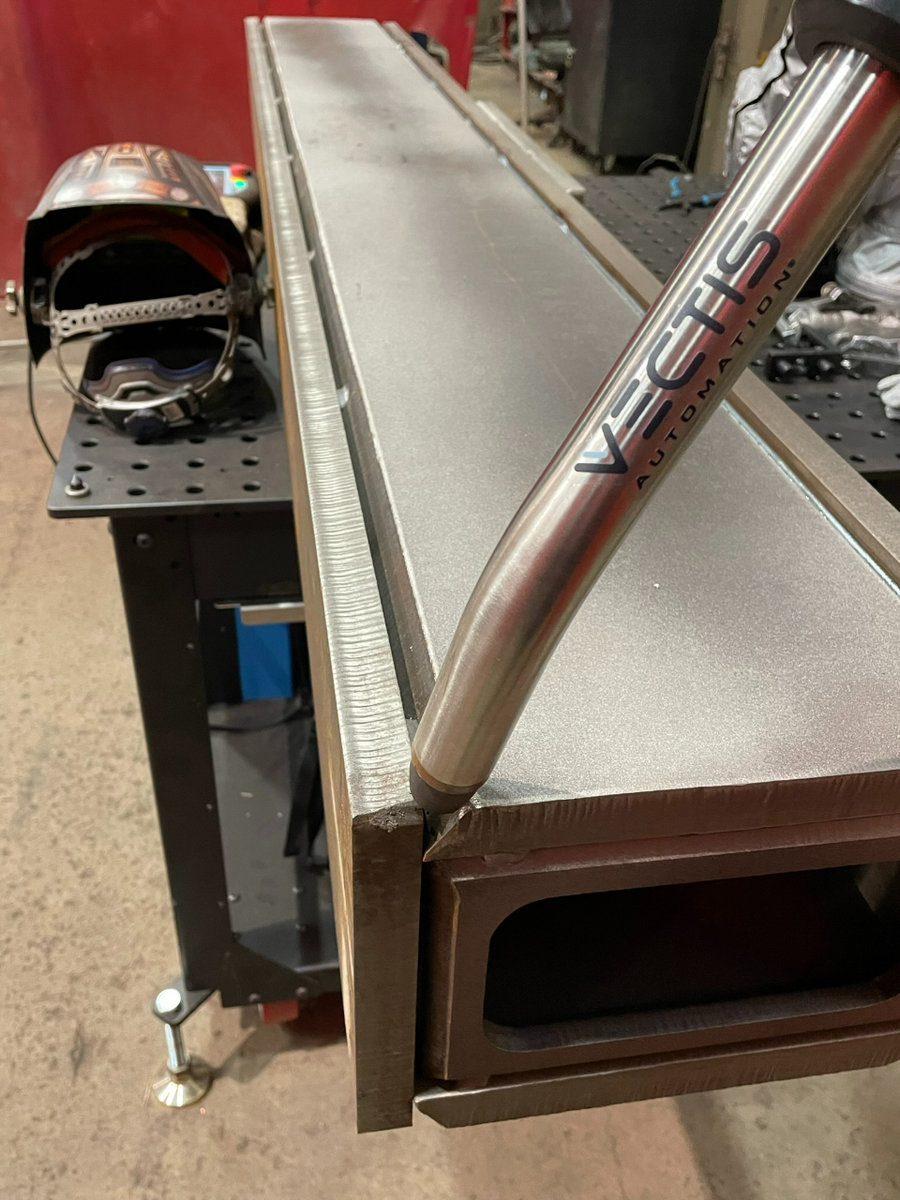 A Vectis brand welding tip attached to an out-of-frame robotic arm prepares to weld two sides of a rectangular prism piece of metal together. 